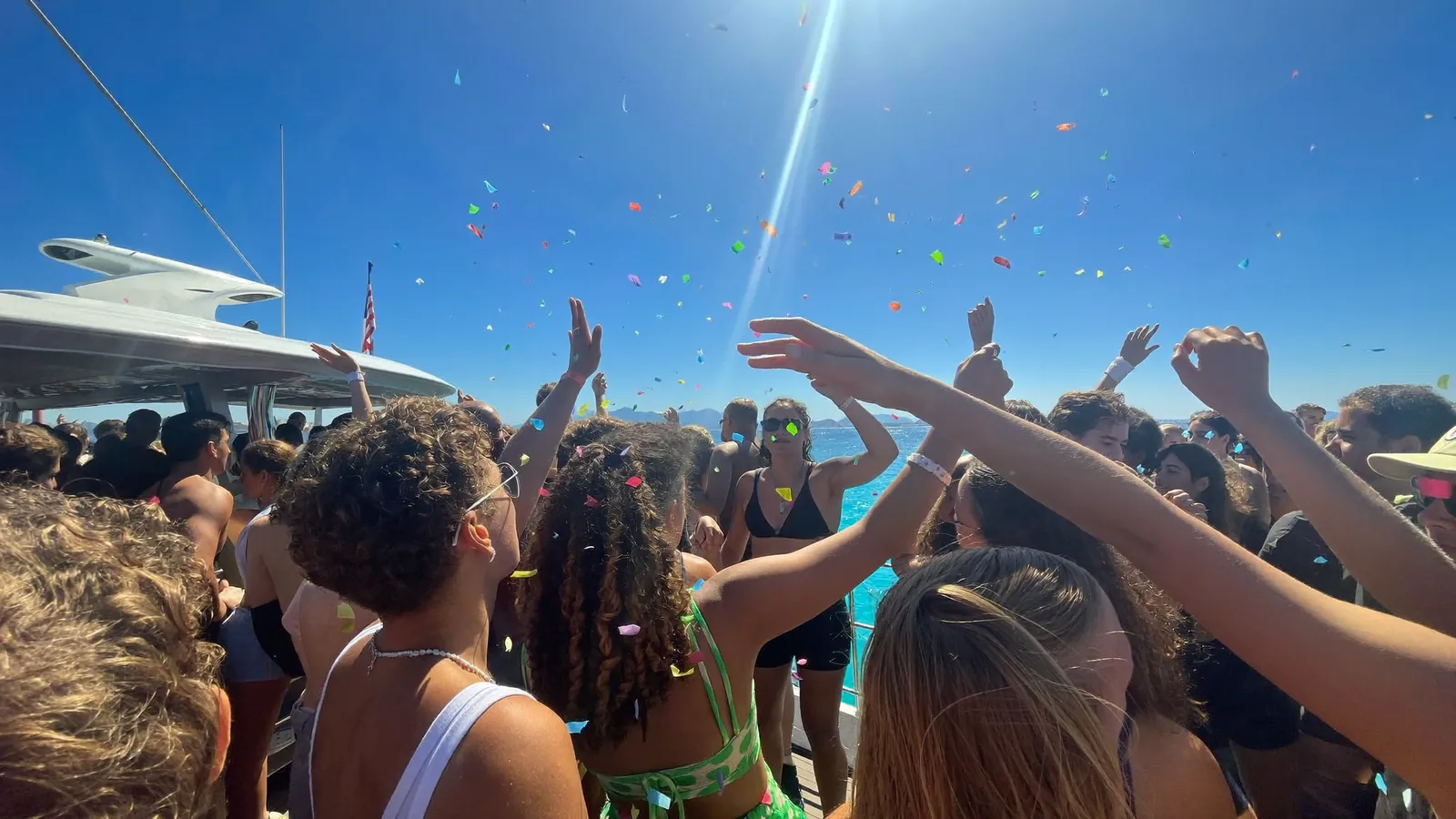 A crowd with their hands in the air and confetti falling on a sunlit boat deck